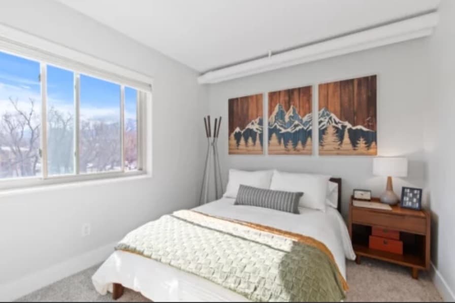 A bright Mountainaire Boulder bedroom with a large window, white walls, a double bed with white bedding and a green throw, a nightstand with a lamp, and triptych mountain artwork above the bed.