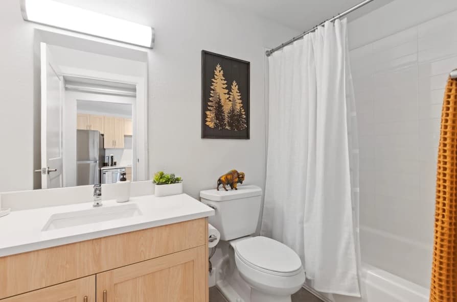 Modern bathroom with a light wood vanity, white countertop, and potted plants creates a relaxing Mountainaire vibe. A toilet with a decorative figure, wall art above, and a shower-tub combo with white curtain and yellow towel complete the look.