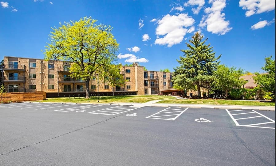 A three-story Mountainaire Boulder apartment building with balconies, surrounded by trees and greenery, stands behind an empty parking lot with several marked accessible parking spaces under a blue sky with scattered clouds.