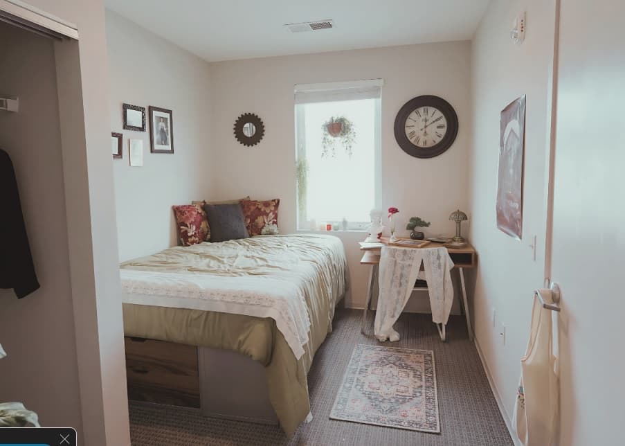A cozy Muse on Main bedroom with a neatly made bed, floral pillows, a small desk with a chair, potted plants, and framed art. Sunlight through the window above the bed creates a warm, inviting atmosphere.