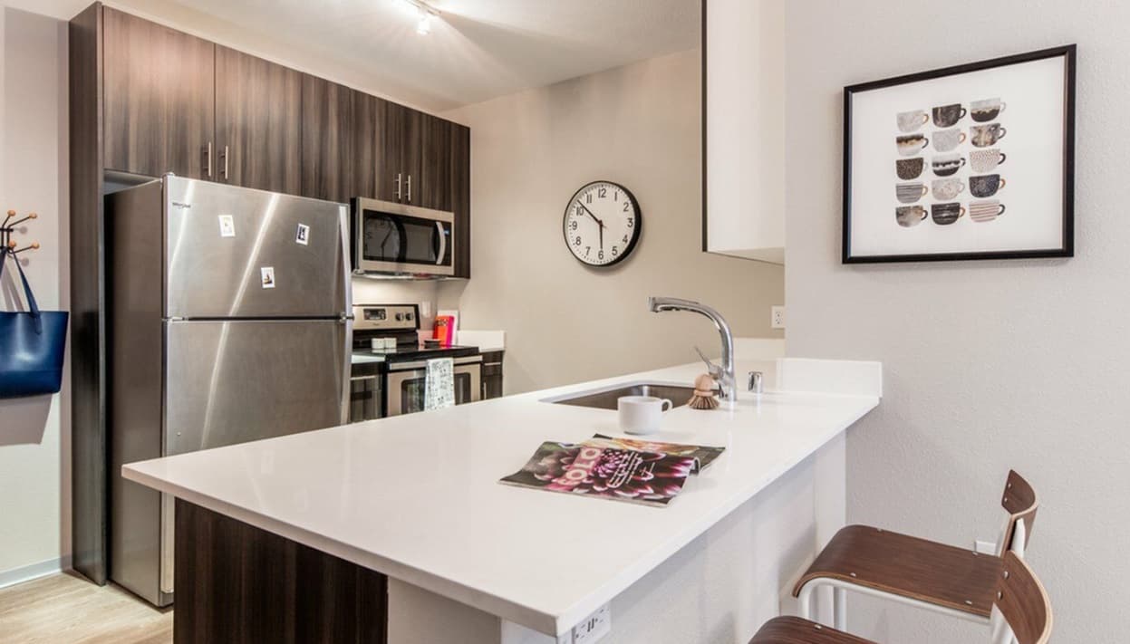 Modern kitchen at Muse on Main with stainless steel appliances, dark cabinets, a white island countertop with two wooden stools, a wall clock, and framed artwork. A mug and a magazine sit on the counter.