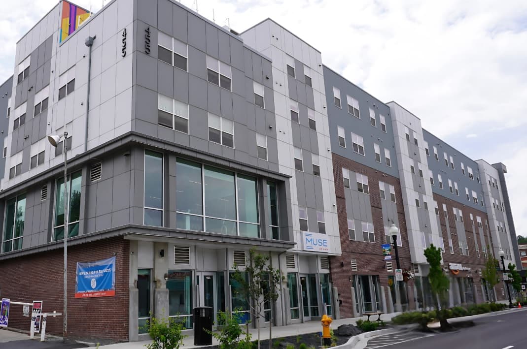 A modern multi-story apartment building with large windows, gray and white paneling, and a brick ground floor features retail spaces and signage, including Muse on Main Apartments, along a city street lined with trees and a fire hydrant.