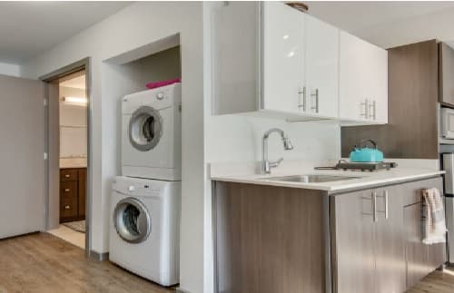 Modern kitchen at Nora apartments with brown cabinets, white countertops, and a turquoise kettle on the stove. A stacked washer and dryer are in a small laundry nook next to an open door leading to a bathroom.