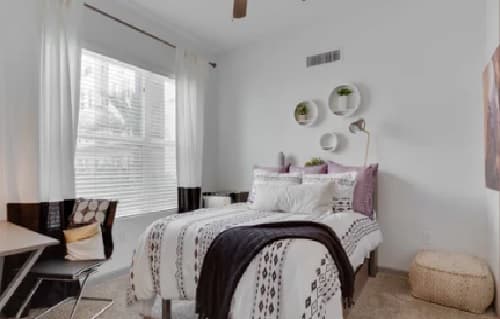 A cozy Nine20 Tempe bedroom features a bed with white and patterned bedding, decorative pillows, and a dark throw blanket. A desk and chair sit by a large window with white curtains. Three round shelves with plants hang above the bed.