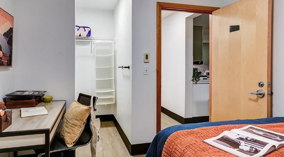 A tidy bedroom in Nordheim Court features a bed, desk, and chair—ideal for UW student apartments. The closet with shelves holds a purple item, while the open door reveals another room. An orange blanket and magazines rest on the bed.