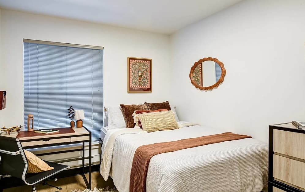 A cozy bedroom in Nordheim Court features a neatly made bed with brown and beige pillows, a rust-colored throw, a desk and chair by the window with blinds—ideal for UW student apartments. A dresser and wall art complete this perfect student housing retreat.