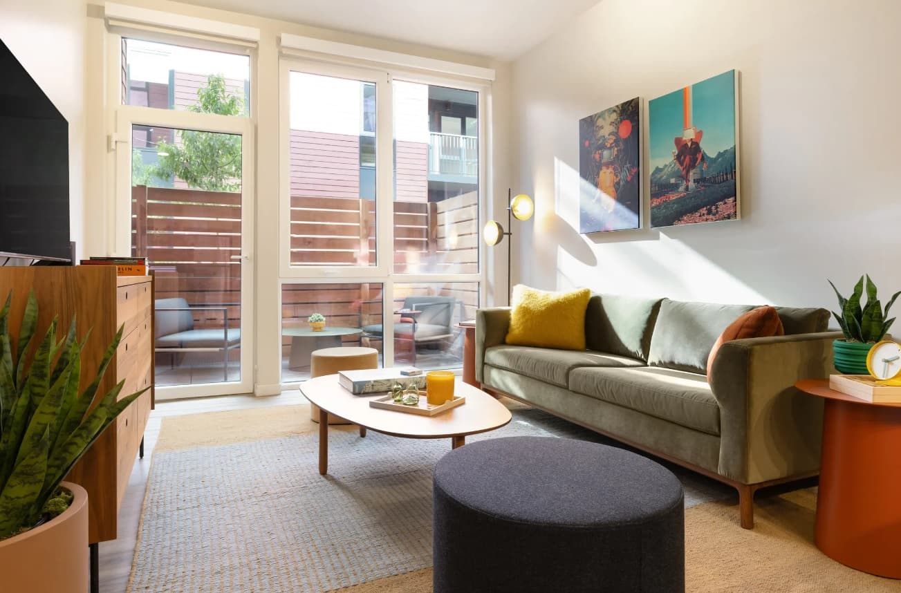A bright, modern living room at Wolf Run Student Housing Apartments Reno features a green sofa, colorful cushions, wall art, round coffee table, potted plant, and floor-to-ceiling windows opening to a small patio with outdoor seating.