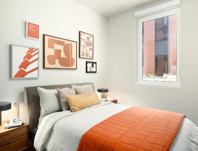 A modern ōLiv Boulder bedroom features a gray bed with white and mustard bedding, an orange throw, and abstract art above. Natural light pours in through a large window. Two nightstands with lamps complete this stylish Boulder retreat.