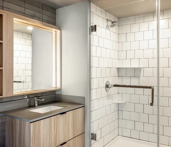 Modern bathroom at ōLiv Boulder Boulder with a wood vanity, rectangular sink, large mirror with built-in lighting, and a walk-in shower featuring white subway tile, glass door, metal grab bar, and built-in shelves.