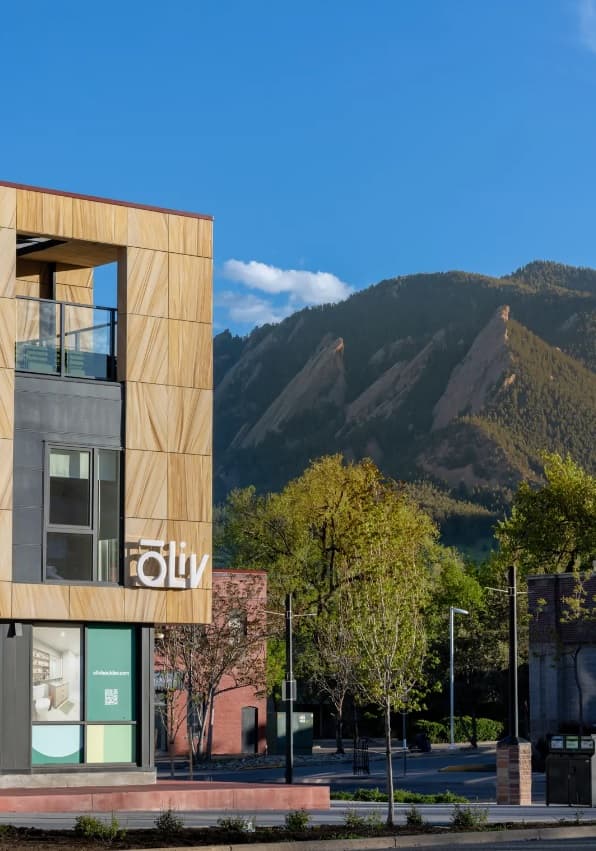 A modern ōLiv Boulder building with a liv sign stands in the foreground, while green trees and rocky, forested mountains rise in the background under a clear blue sky.
