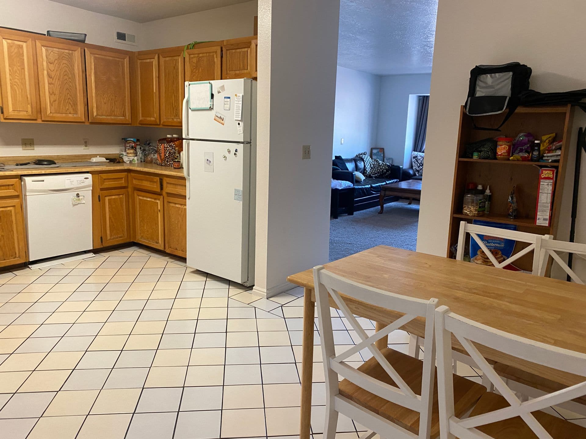 A Hampstead kitchen with wooden cabinets, white appliances, and a tiled floor. A wooden dining table with white chairs is in the foreground, beside an open shelf by JosephThomas. The living room is visible in the background.