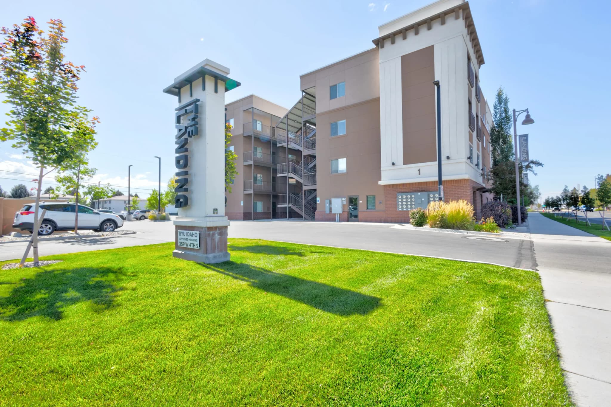 A modern, four-story apartment building with tan and white exterior walls stands behind a green lawn with a tall, vertical sign reading INDIGO near The Landing Rexburg driveway and parked cars.