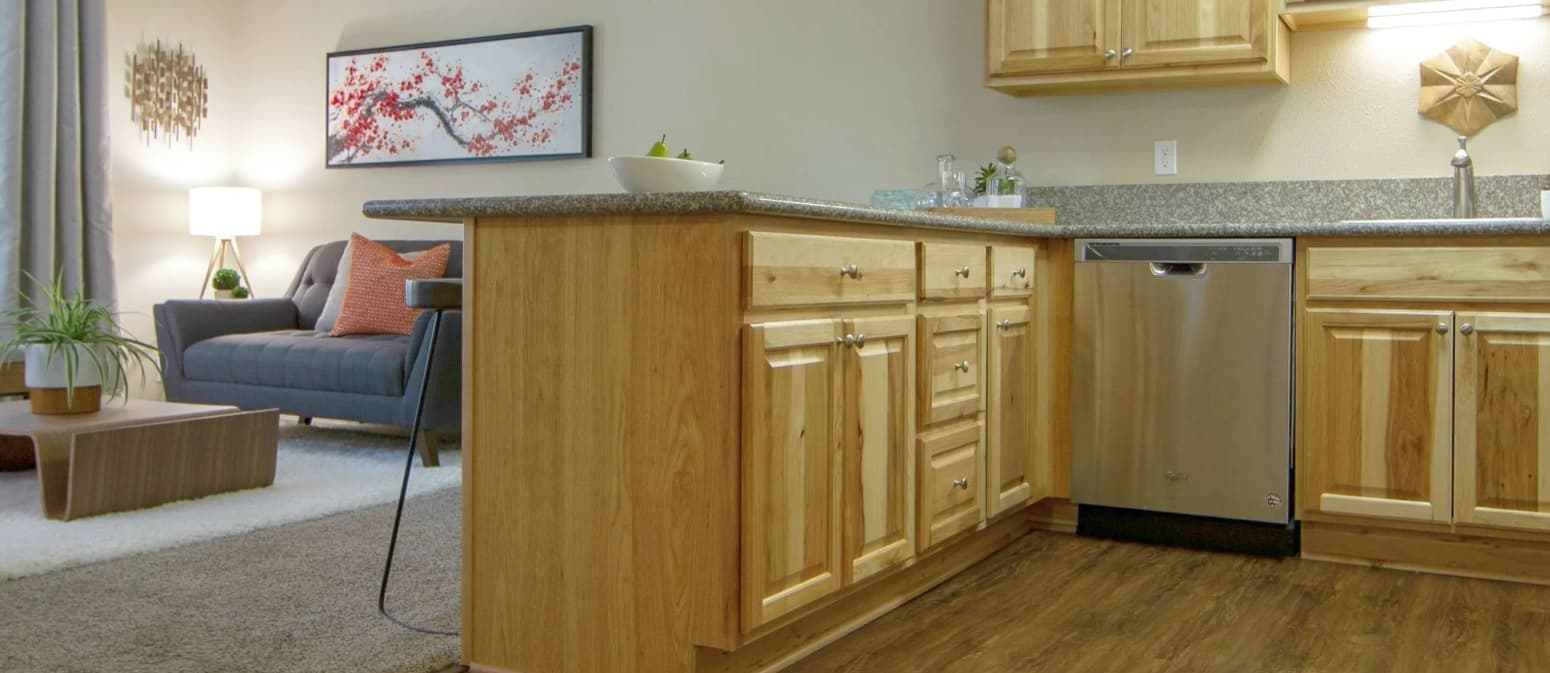 A modern kitchen with light wood cabinets and a gray countertop at Oak Vale Apartments, adjoining a cozy living area with a gray sofa, white rug, potted plant, and wall art featuring red and black branches.