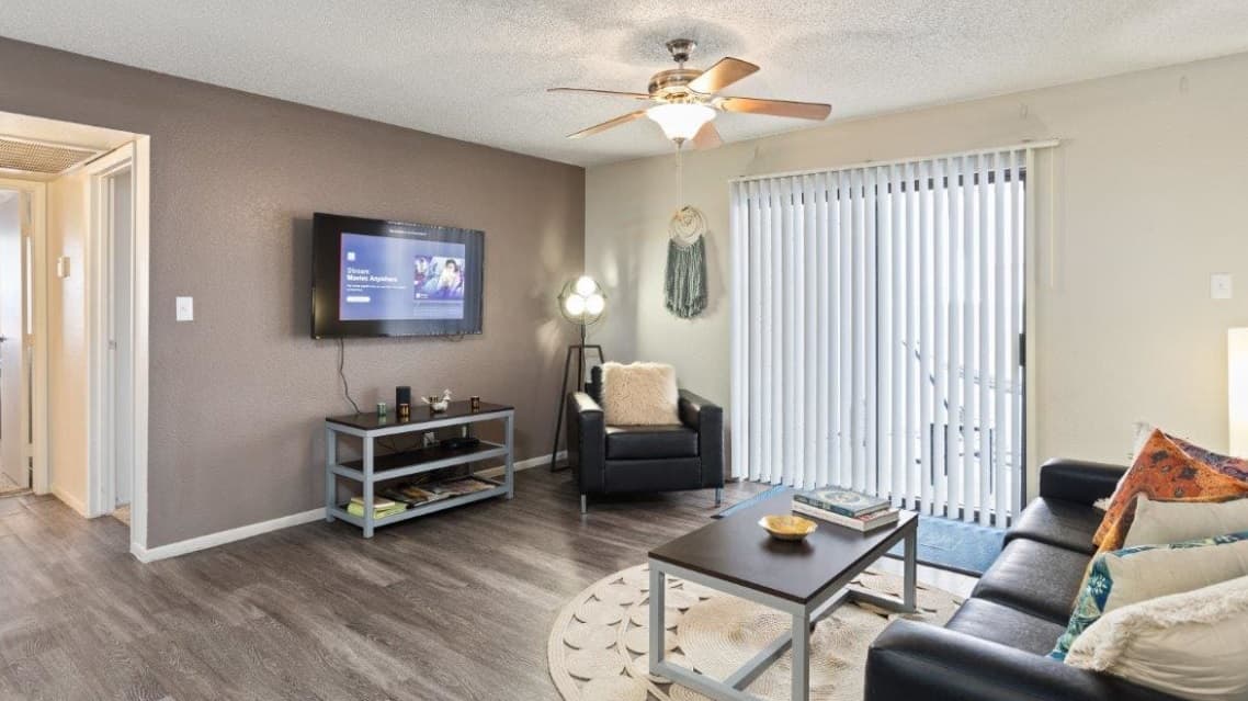 A modern living room at Paseo On University in Tempe features a mounted TV, black armchair and sofa, coffee tables, throw pillows, ceiling fan, and sliding glass doors with vertical blinds letting in natural light—perfect for student housing.