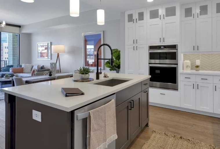 Modern kitchen with white cabinets, built-in oven, and a gray island with sink and towel bar—perfect for luxury apartments. An open living area with sofa and large window creates a welcoming atmosphere in Pullman-inspired style. Warm, neutral tones throughout.