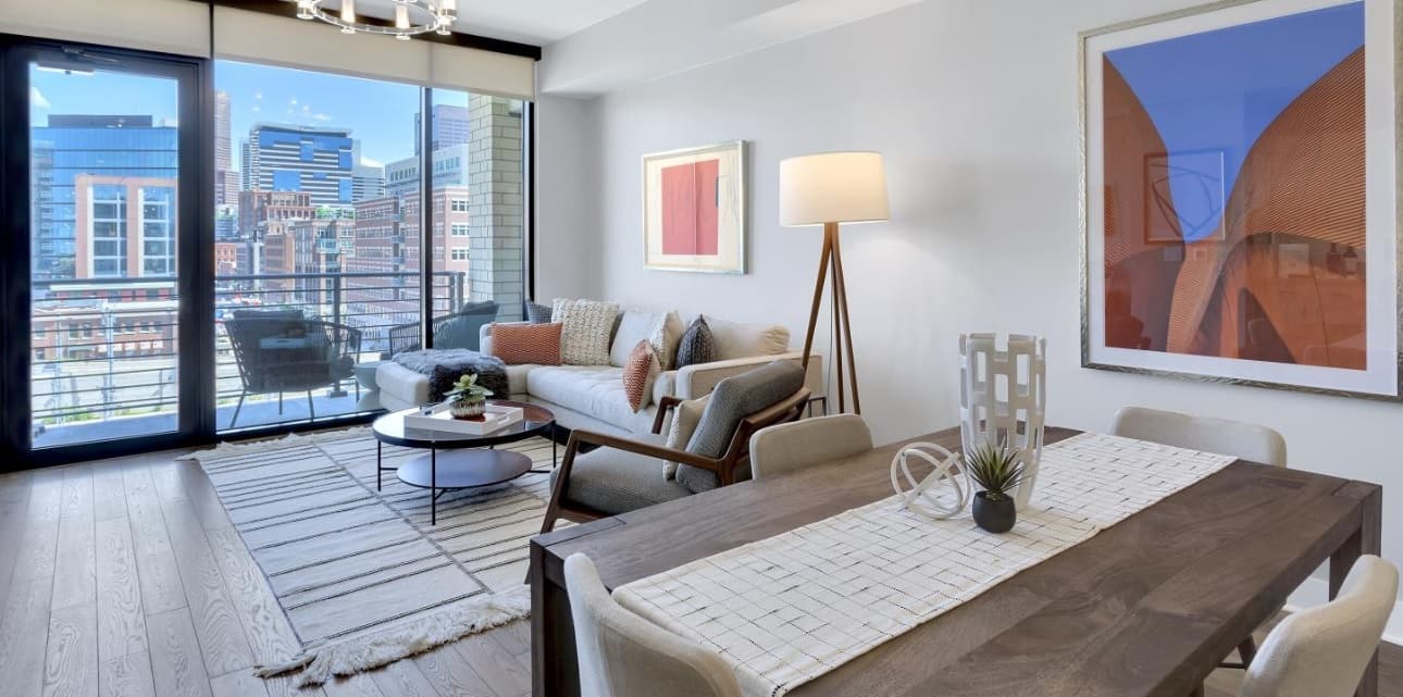 Modern living and dining area in Pullman luxury apartments with light wood floors, neutral decor, a beige sofa, framed art, floor lamp, and large windows revealing a city skyline and balcony. Dining table is set for four.