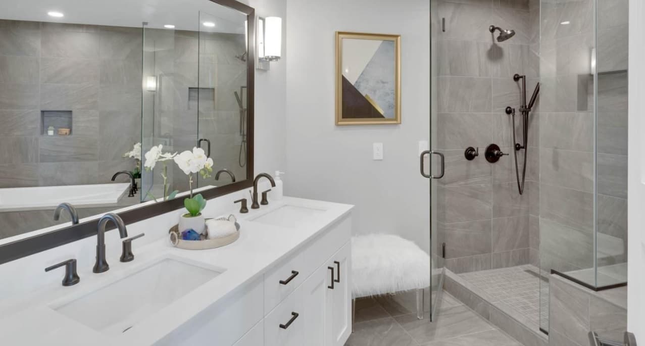 Modern bathroom at Pullman Luxury Apartments with a double-sink vanity, large mirror, flower arrangement, glass-enclosed shower with black fixtures, fluffy white stool, and abstract wall art. The space features neutral colors and sleek finishes.
