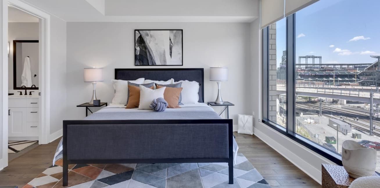 A modern bedroom in Pullman Luxury Apartments features a black-framed bed with white bedding and brown accent pillows. Large windows reveal a city view with buildings and a stadium, while abstract art hangs above the headboard.