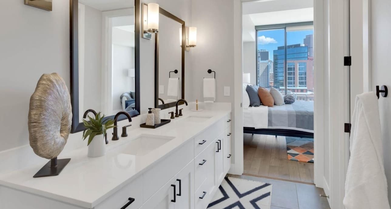 Modern bathroom with double sinks, large mirrors, black fixtures, and white cabinets in Pullman Luxury Apartments. A bedroom with large windows and a city view is visible through an open doorway. Decorative accents and a towel add style.