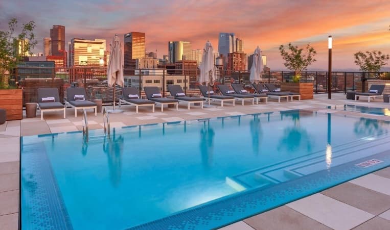 A rooftop pool with lounge chairs and umbrellas at Pullman Luxury Apartments overlooks a city skyline at sunset, with colorful clouds in the sky and tall buildings in the background.