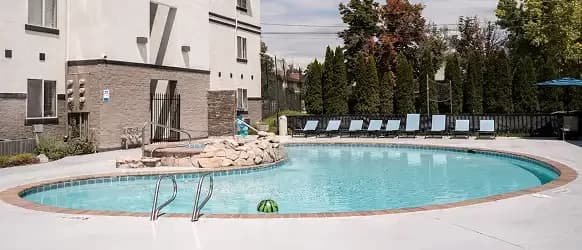 A small outdoor swimming pool with a curved shape at Crestwood Provo, surrounded by lounge chairs and umbrellas. There’s a shallow entry with rocks and a handrail, set next to a building and tall trees in the background.