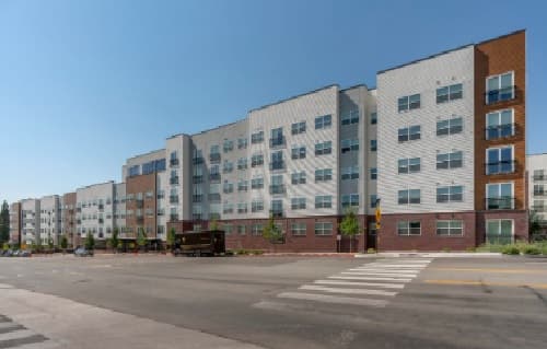 A large modern apartment building, Park Place at Reno Reno, features five stories with alternating white and brown panels, many windows, and balconies. It is situated along a wide street with a pedestrian crosswalk in front.