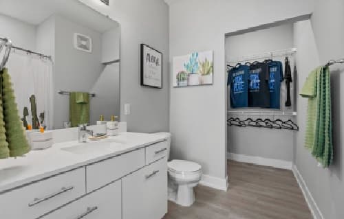 Modern bathroom at Park Place at Reno Reno with a white vanity, sink, and toilet. Green towels hang on racks. Wall art and toiletries sit on the counter, with an open closet in the background featuring neatly hung clothes and empty hangers.