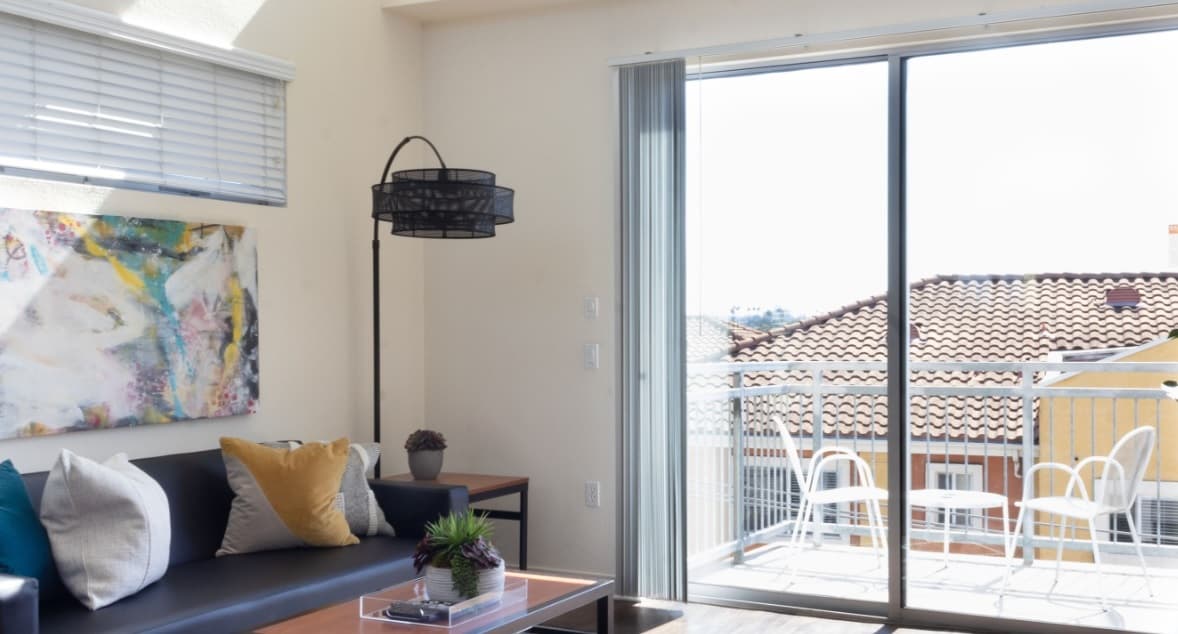 A bright living room at Paseo Place San Diego features a black sofa, colorful pillows, abstract wall art, and sliding glass doors that open to a sunny balcony with two white chairs and a table.
