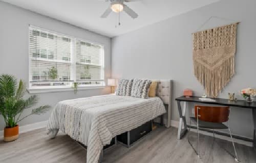 A modern, tidy bedroom at Park Place at Reno Reno features a neatly made bed, a potted plant, a desk with a chair, and a decorative macrame wall hanging. Sunlight streams in from a large window, brightening the room.