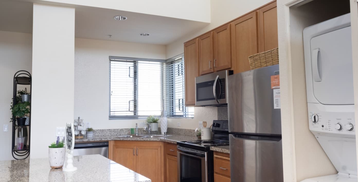 Modern kitchen at Paseo Place San Diego with wood cabinets, granite countertops, stainless steel appliances, a corner sink under two windows with blinds, and a stacked washer and dryer in a nearby closet. Decorative plants and kitchen items are visible.