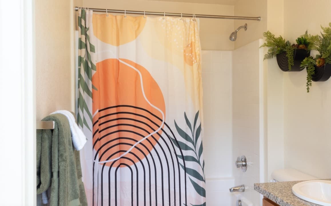 A bathroom at Paseo Place San Diego features a colorful shower curtain with abstract sun and plant designs, a towel on a rack, a wall-mounted plant, and part of the sink and countertop in view.