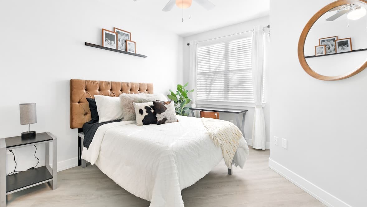Bright bedroom at Park Place Tempe with a tan tufted headboard, white bedding, black-and-white accent pillows, nightstand, lamp, wall art, plant, a desk by the window, and a round mirror on the wall.