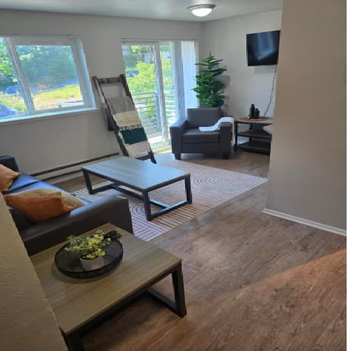 Modern living room in Pacific Sunrise with wood flooring, a brown sofa, coffee table, armchair, wall-mounted TV, potted plant, and large window letting in natural light. A ladder shelf with blankets stands near the window. Ideal for U-District Student Housing.
