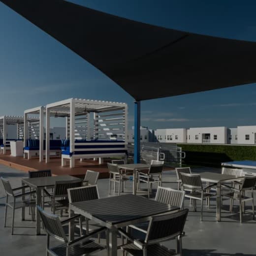 Outdoor patio with metal tables and chairs, shaded by a large black canopy. In the background, there are white cabanas with blue cushions and white modern buildings under a clear sky.