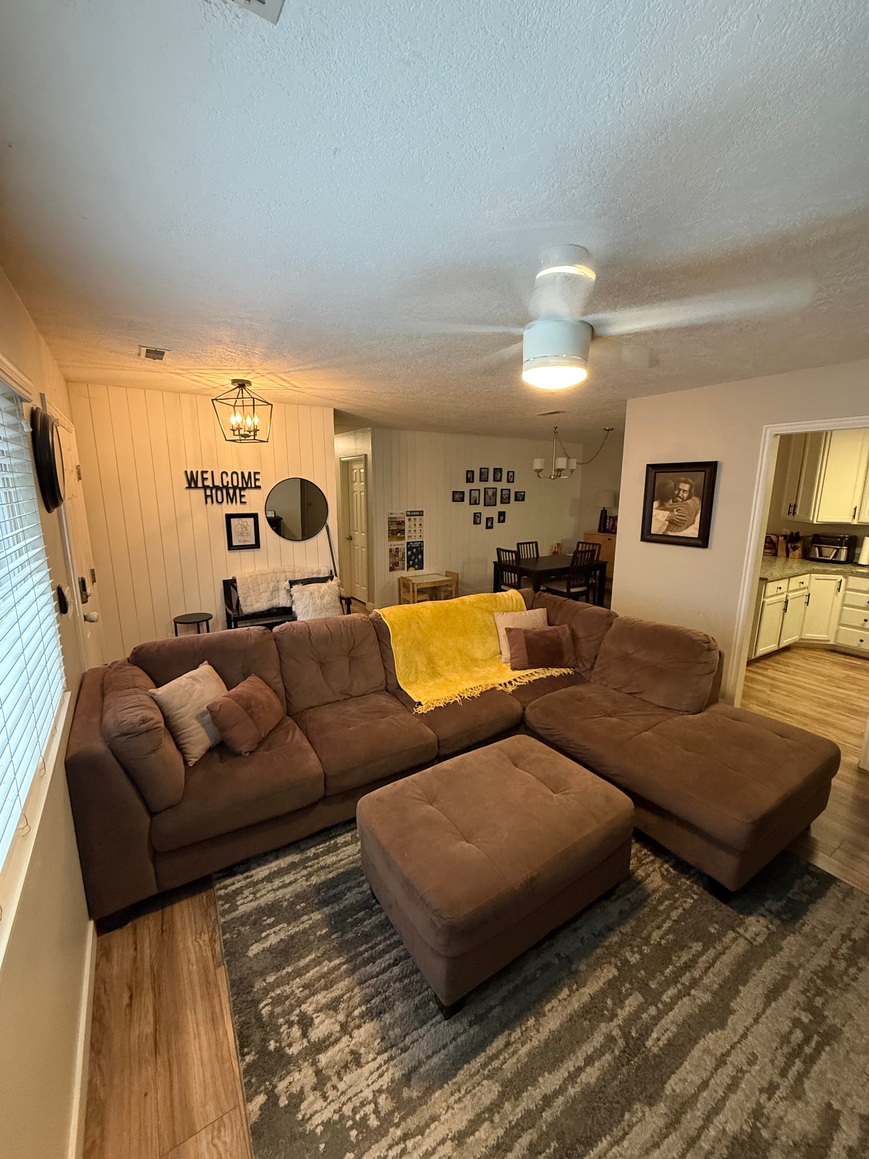 A cozy apartment living room at 282 W 960 S features a brown sectional sofa, tan and white pillows, a yellow blanket, matching ottoman, and gray rug. Wall decor says WELCOME HOME. The dining area and kitchen are visible in the background.
