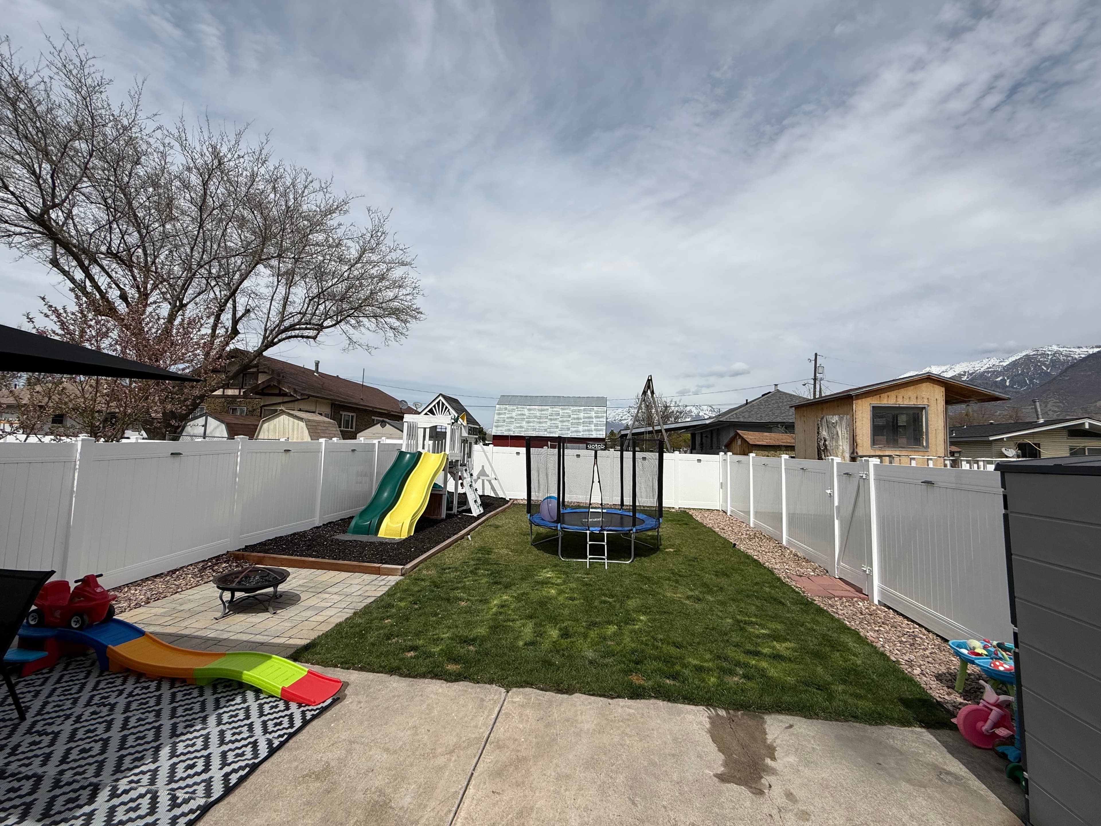 A fenced backyard at APT C, 282 W 960 S features a playground set with a yellow slide, small trampoline, playhouse, and toddler slide on a mat. The yard has grass and concrete, surrounded by nearby houses and trees.