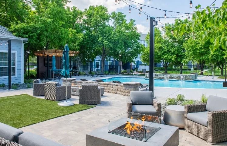 Outdoor patio area at Campus Crossings at Ram's Pointe Fort Collins features modern wicker seating, a fire pit, string lights, and a swimming pool surrounded by trees and lush landscaping. A shaded pergola and blue umbrella complete the inviting space.