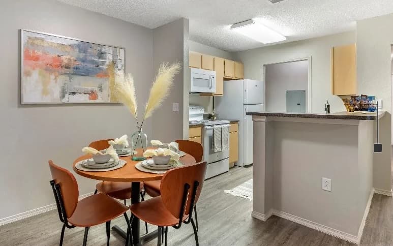 A modern apartment kitchen at Campus Crossings at Ram's Pointe Fort Collins features light wood cabinets, white appliances, and a breakfast bar. A small round dining table is set for two with brown chairs, decorative vases, and abstract art on the wall.
