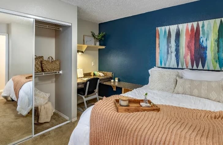 A cozy bedroom at Campus Crossings at Ram's Pointe Fort Collins features a blue accent wall, white and beige bedding, a wooden tray, desk with chair and lamp, woven baskets in an open closet, and colorful abstract art above the bed.