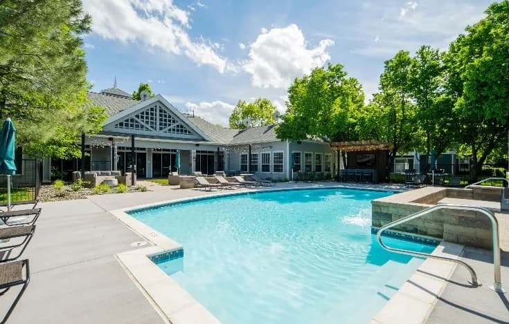 Outdoor swimming pool with clear blue water, surrounded by lounge chairs, trees, and the modern Campus Crossings at Ram's Pointe Fort Collins clubhouse under a sunny sky with scattered clouds.