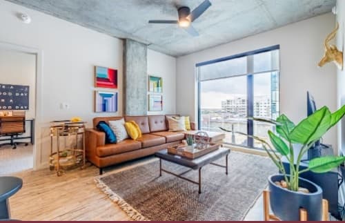 A modern living room at Rev Apartments with a brown leather sofa, colorful cushions, wall art, a large plant, coffee table, floor rug, and floor-to-ceiling windows overlooking city buildings—ideal for Northgate student living. A small office is visible through an open door.