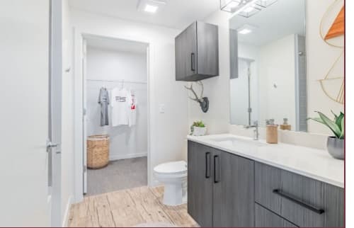 Modern bathroom with gray cabinets, a large mirror, white countertop, toilet, and light wood flooring at Rev Northgate. An adjacent walk-in closet is visible, containing hanging clothes and a woven laundry basket—ideal for student living.