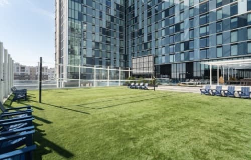A grassy outdoor rooftop area at Rev Northgate features a volleyball court, blue chairs, loungers, and views of a high-rise glass building. This student accommodation space is enclosed with glass panels and brightly lit by sunlight.