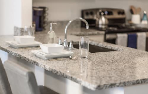 A modern kitchen at Sierra Student Apartments features a granite countertop, two place settings with square white bowls and plates, drinking glasses, a soap dispenser, and a stainless steel sink. In the background are a stove and kettle.