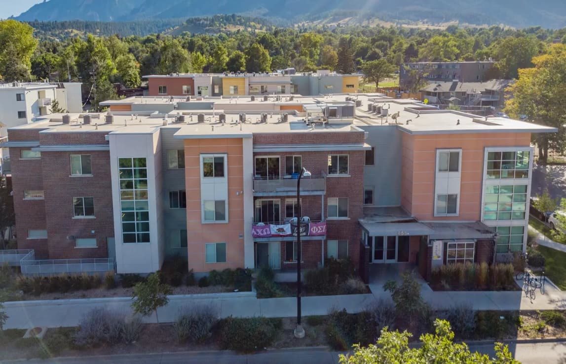 Aerial view of the modern three-story Saga Boulder apartment building with large windows and balconies, set against a backdrop of trees and distant mountains on a sunny day.