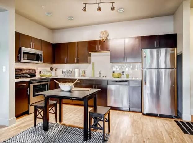 Modern kitchen with dark wood cabinets and stainless steel appliances features a Saga Boulder dining table with two benches on a striped rug. Light wood floors, minimalist decor, and bright lighting complete the look.