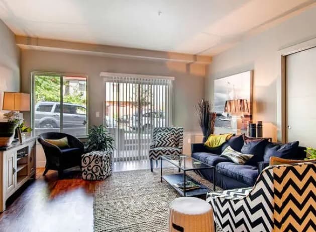 A cozy living room features a dark blue Saga Boulder sofa, patterned armchairs, a glass coffee table, a rug, indoor plants, and large windows with vertical blinds letting in natural light and showing a view of a parked car outside.