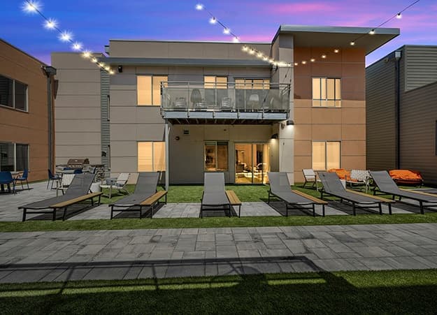 Modern outdoor lounge area with several reclining chairs on artificial grass, illuminated by string lights, beside the Saga Boulder Boulder—a two-story building with large windows and a balcony at dusk.