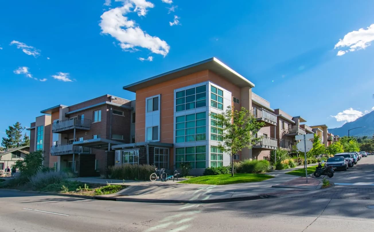 Modern three-story apartment building with large windows and balconies, situated on a corner lot under a clear blue sky in the heart of Saga Boulder Boulder. Trees, bikes, and parked cars line the street in front.