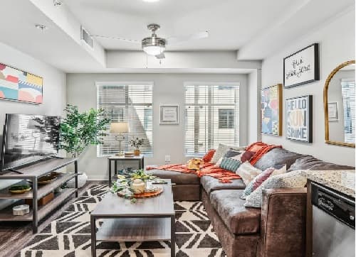 Bright living room at The Standard at Berkeley with a brown sectional sofa, colorful cushions, wall art, patterned rug, TV on a stand, coffee table with decor, and large windows letting in natural light.